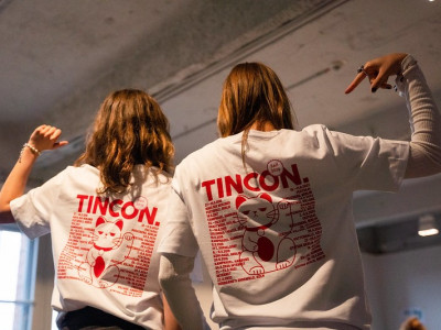 Zwei junge Menschen stehen nebeneinander mit dem Rücken zur Kamera und tragen weiße T-Shirts mit rotem TINCON-Logo und einem großen Druck einer winkenden Glückskatze. Sie zeigen auf ihre T-Shirts.