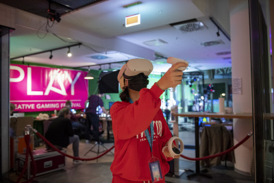 Foto vom PLAY Festival. Im Vordergrund zu sehen ist eine Person in einem roten Pulli, welche eine VR-Brille trägt und in beiden Händen jeweils einen Controller hält und dabei gerade den rechten Arm für eine Aktion hebt. Im Hintergrund hängt ein Banner mit 
