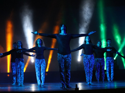 Sieben dunkel gekleidete Personen auf Bühne mit bunten Lichtern während einer Tanzperformance.