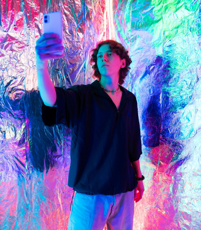 Junger Mann macht Selfie vor einer mit Alufolie bespannten Wand die bunt angestrahlt ist