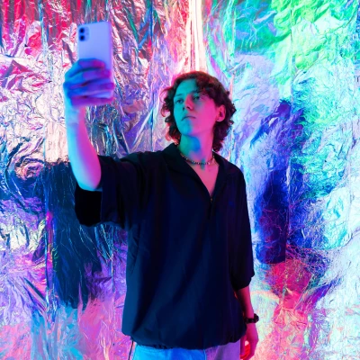 Junger Mann macht Selfie vor einer mit Alufolie bespannten Wand die bunt angestrahlt ist