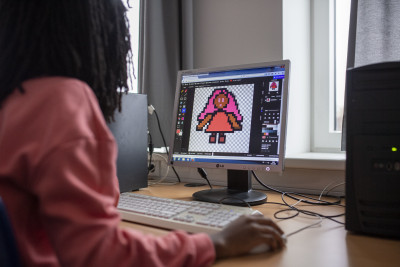 Zu sehen ist eine junge Person, mit dunklen, schulterlangen Haaren mit, in lachsfarbenem Langarmoberteil, die an einem Workshop teilnimmt und gerade vor einem PC-Bildschirm sitzt und eine Grafik in Pixel-Optik erstellt.