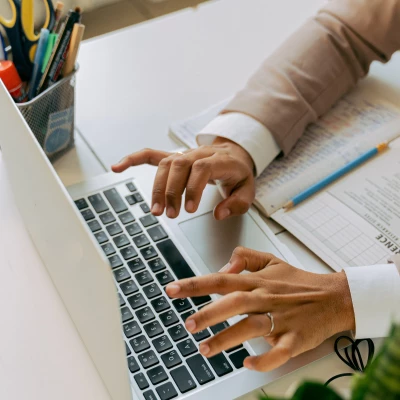 Zu sehen sind Hände einer Person, die am Laptop etwas tippt. Vor ihr liegt ein Notizheft mit einem Bleistift und neben dem Laptop eine Behälter mit verschiedenen Utensilien, wie Stifte und Schere.