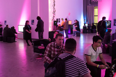 Schnappschuss aus der Ausstellung vom PLAY – Creative Gaming Festival. Zu sehen ist ein in rosa Licht getauchter Raum, mit mehreren ausgestellten Spielen, an denen Menschen sitzen oder stehen und spielen und sich unterhalten.