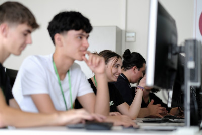 Zu sehen sind fünf Jugendliche in einem Klassenraum, die vor Bildschirmen sitzen. Eine Person tippt, die daneben schaut nach vorn, die dritte Person lacht und die letzten beiden unterhalten sich.