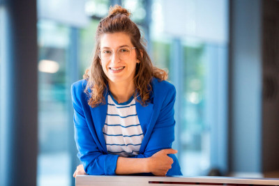 Foto von Dr. Julia Köhler, der Projektleiterin von data driven future. Lächelnd, mit schulterlangen, rot-braunen, halb-offenen Haaren, in blauem Jackett und blau-weiß gestreiftem Oberteil vor einer Fensterfront.