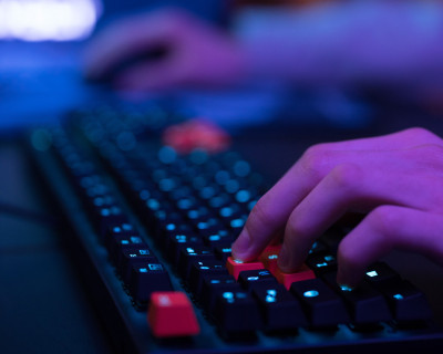Zu sehen ist eine schwarze Tastatur und verschwommen im Hintergrund eine PC-Maus, die jemand bedient.
