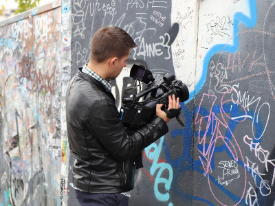 Mensch in schwarzer Jacke mit großer Videokamera in der Hand filmt eine Graffiti-Wand.