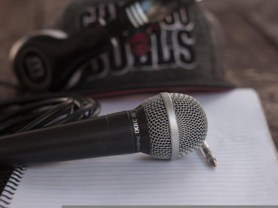 Mikrofon liegt auf einem Schreibblock. Daneben ein Kugelschreiber. Im Hintergrund Kopfhörer und Cap.