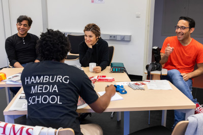 Fotos von vier Personen, die bei einem Tag der Offenen Tür der Hamburg Media School vor Ort waren. Eine Person ist in der Rückenansicht zu sehen und trägt ein dunkelblaues T-Shirt mit dem Aufdruck 