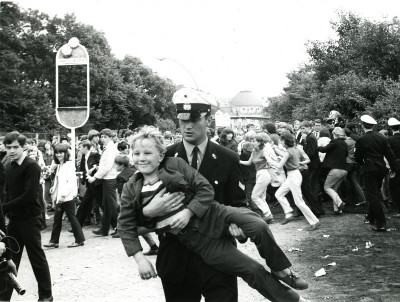 Schwarz-Weiß-Foto, Polizist trägt lachendes Kind weg, im Hintegrund eine demonstrierende Menge