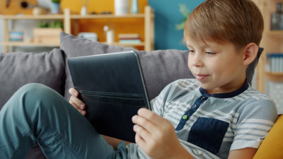 Kind sitzt gemütlich auf dem Sofa und schaut auf ein Tablet