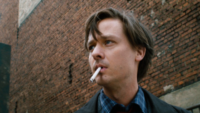 Filmstill, Junger Mann in Nahaufnahme mit Zigarette im Mund vor Backsteinmauer schaut links aus dem Bild