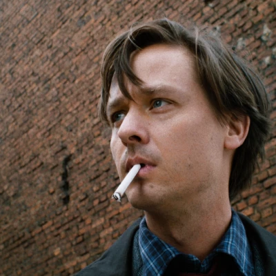 Filmstill, Junger Mann in Nahaufnahme mit Zigarette im Mund vor Backsteinmauer schaut links aus dem Bild