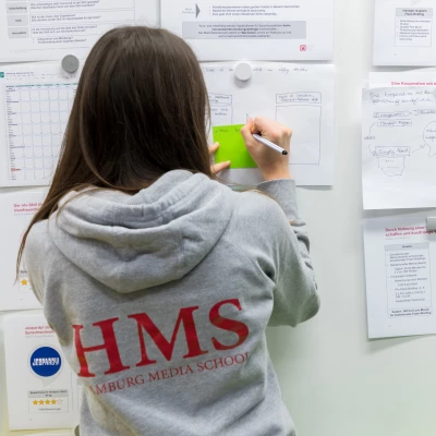 Verschiedene Dokumente mit bunten Magneten an einer weißen Wand befestigt. Davor steht eine Person mit langen, braunen Haaren, in einem Hamburg Media School Kapuzenpulli und schreibt etwas auf einen grünen Notizzettel.