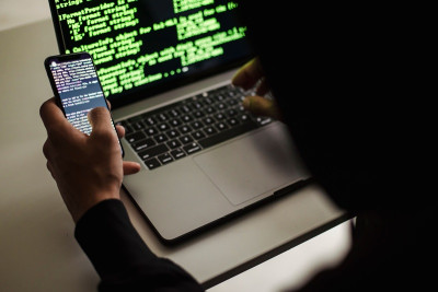 Mensch vor Laptop und Handy, auf denen Computercode zu sehen ist